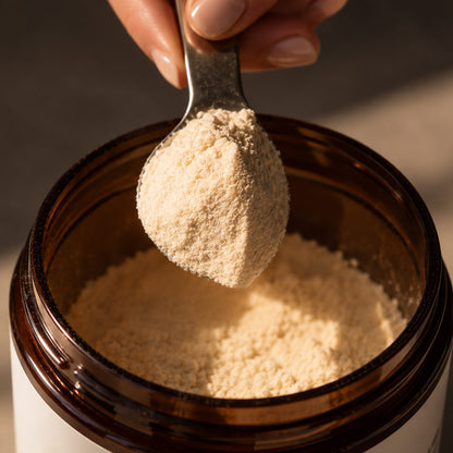 A close-up of a hand holding a metal spoon filled with collagen powder above an amber glass jar containing more of the same powder.