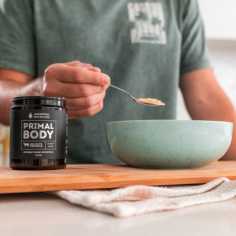 A person in a green t-shirt holds a spoon over a bowl while a jar labeled PRIMAL BODY by ANCESTRAL NUTRITION sits on a wooden board in the foreground.