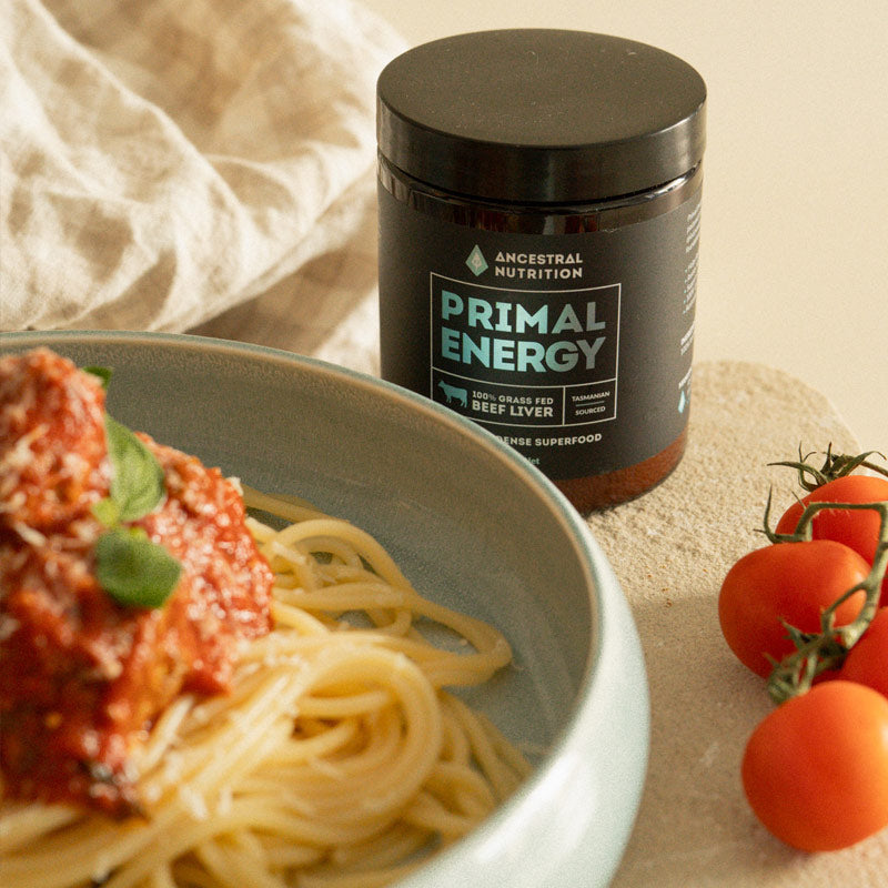 A jar of Ancestral Nutrition Primal Energy beef liver superfood placed next to a bowl of spaghetti with meat sauce and tomatoes on a beige tabletop.
