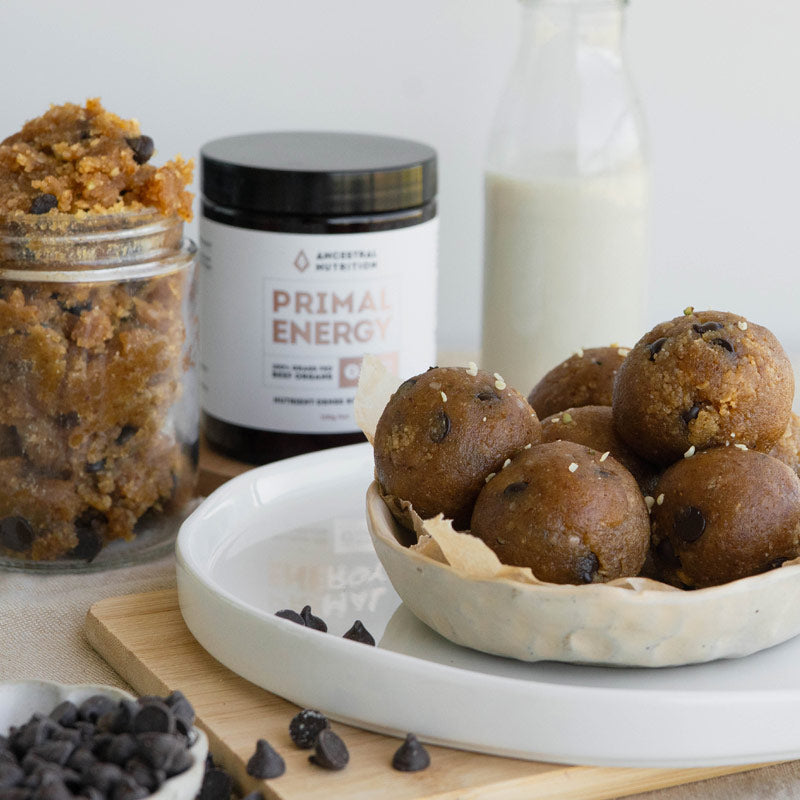 A styled food product shot showing several chocolate chip energy balls in a ceramic bowl on a white plate, with a jar of cookie dough and a bottle of milk in the background. A jar label reading 'Ancestral Nutrition Primal Energy' is visible.