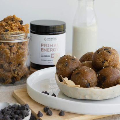 A styled food product shot showing several chocolate chip energy balls in a ceramic bowl on a white plate, with a jar of cookie dough and a bottle of milk in the background. A jar label reading 'Ancestral Nutrition Primal Energy' is visible.