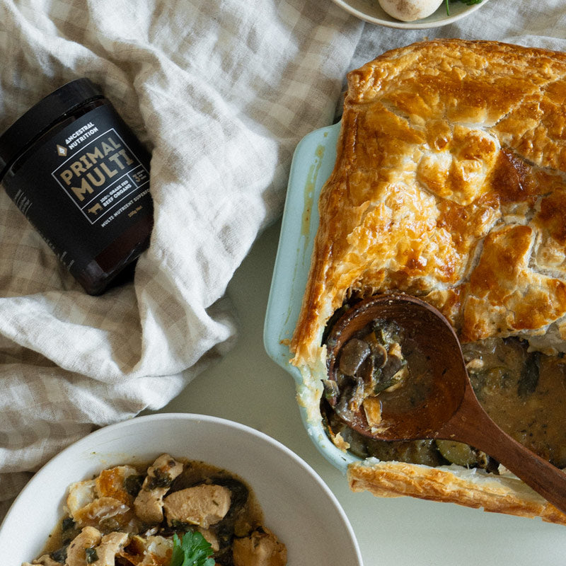 A baked chicken and vegetable pie in a light blue dish with a wooden spoon, styled on a white surface with a bowl of food, a black jar labeled Primal Multi, and a beige checkered cloth.